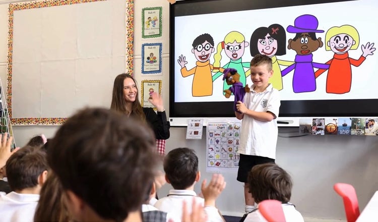 A boy standing in front of his class with a purple Pete Persistence puppet, assisting his teacher, who is sitting to his right, with a lesson on the 5