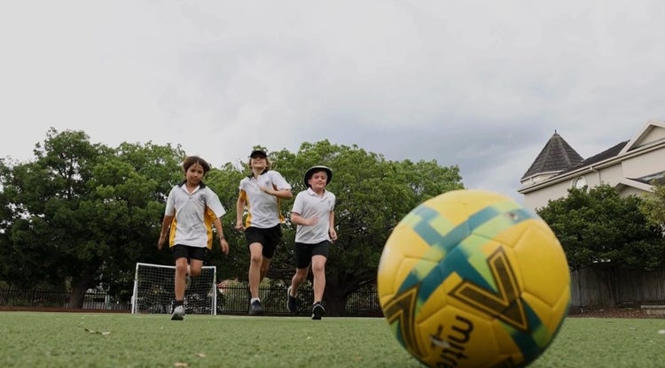3 students running across a soccer field towards a yellow and green soccer ball.